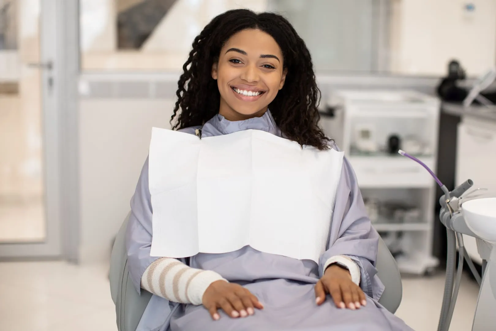 Smiling woman in dental chair wears a dental bib and has bandages on forearms in the office.