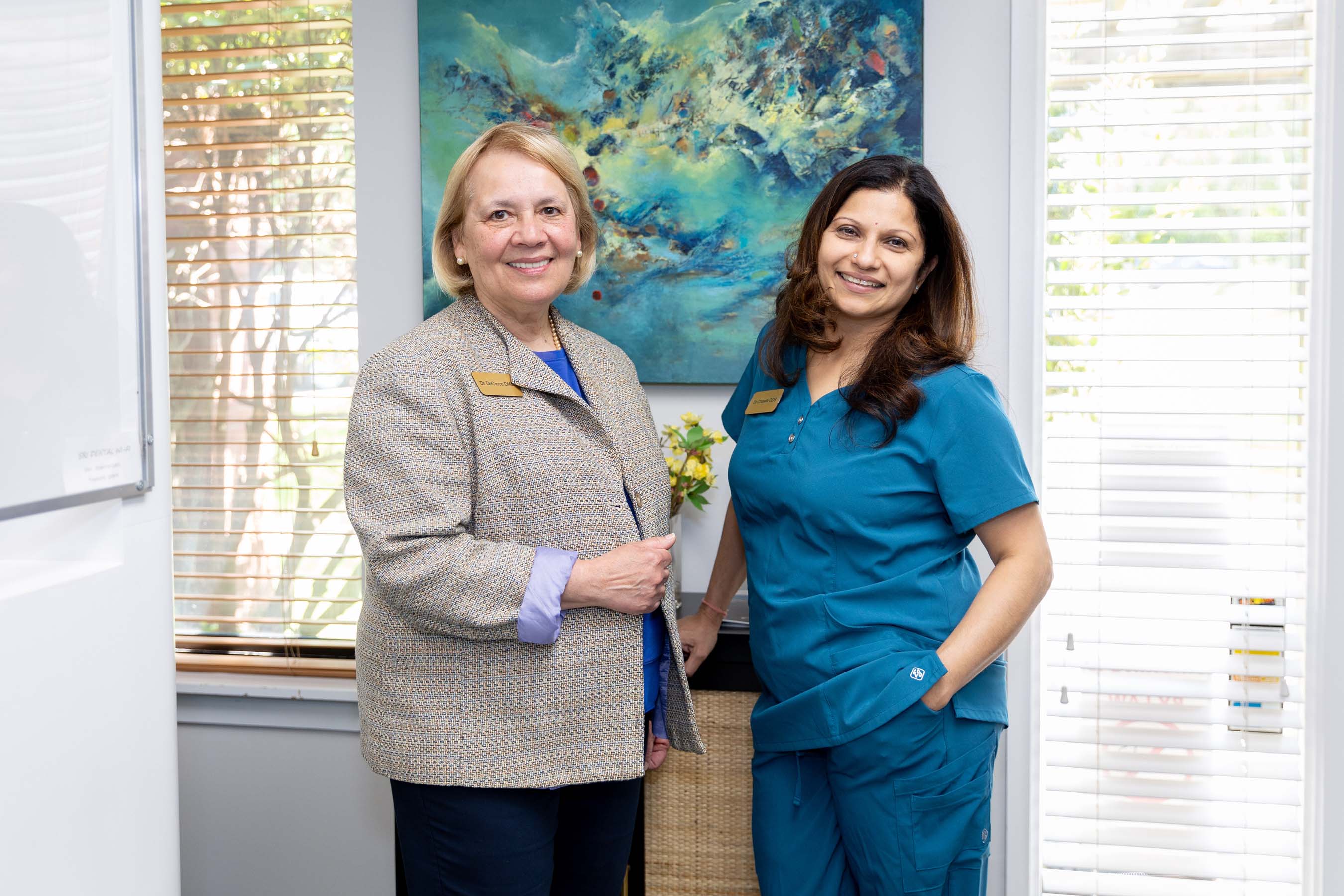 Two smiling women indoors; one in a beige jacket, the other in blue scrubs, with abstract art behind them.