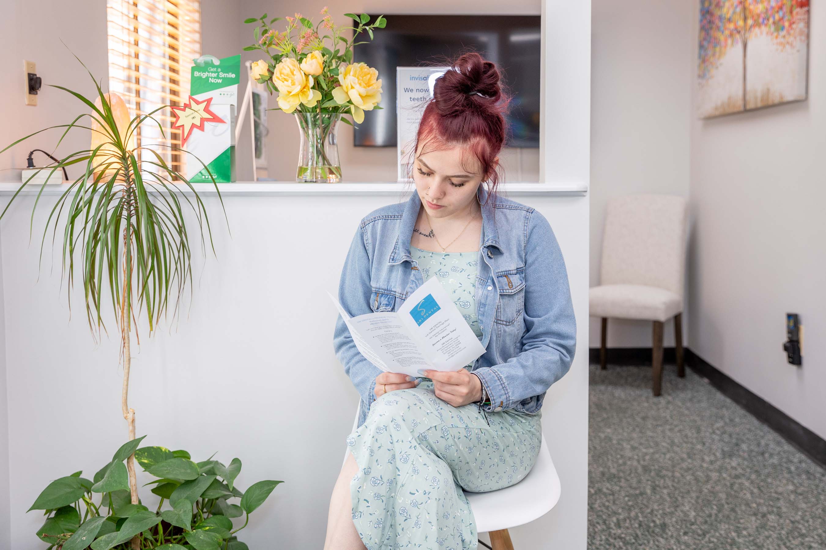 Woman with red hair in a denim jacket reading a brochure in a bright waiting room with plants.