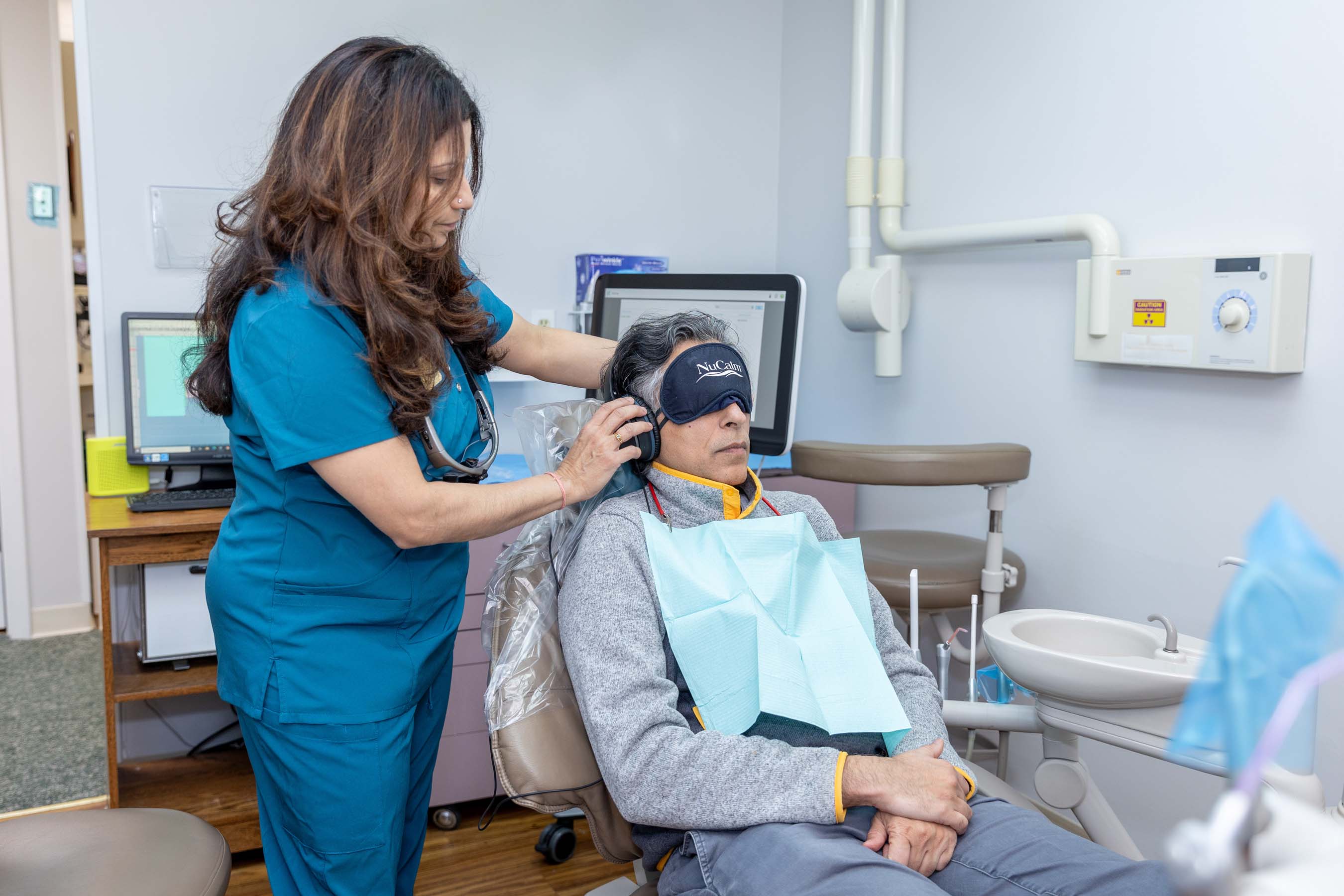 Dental professional fitting headphones on seated patient wearing an eye mask and dental bib.