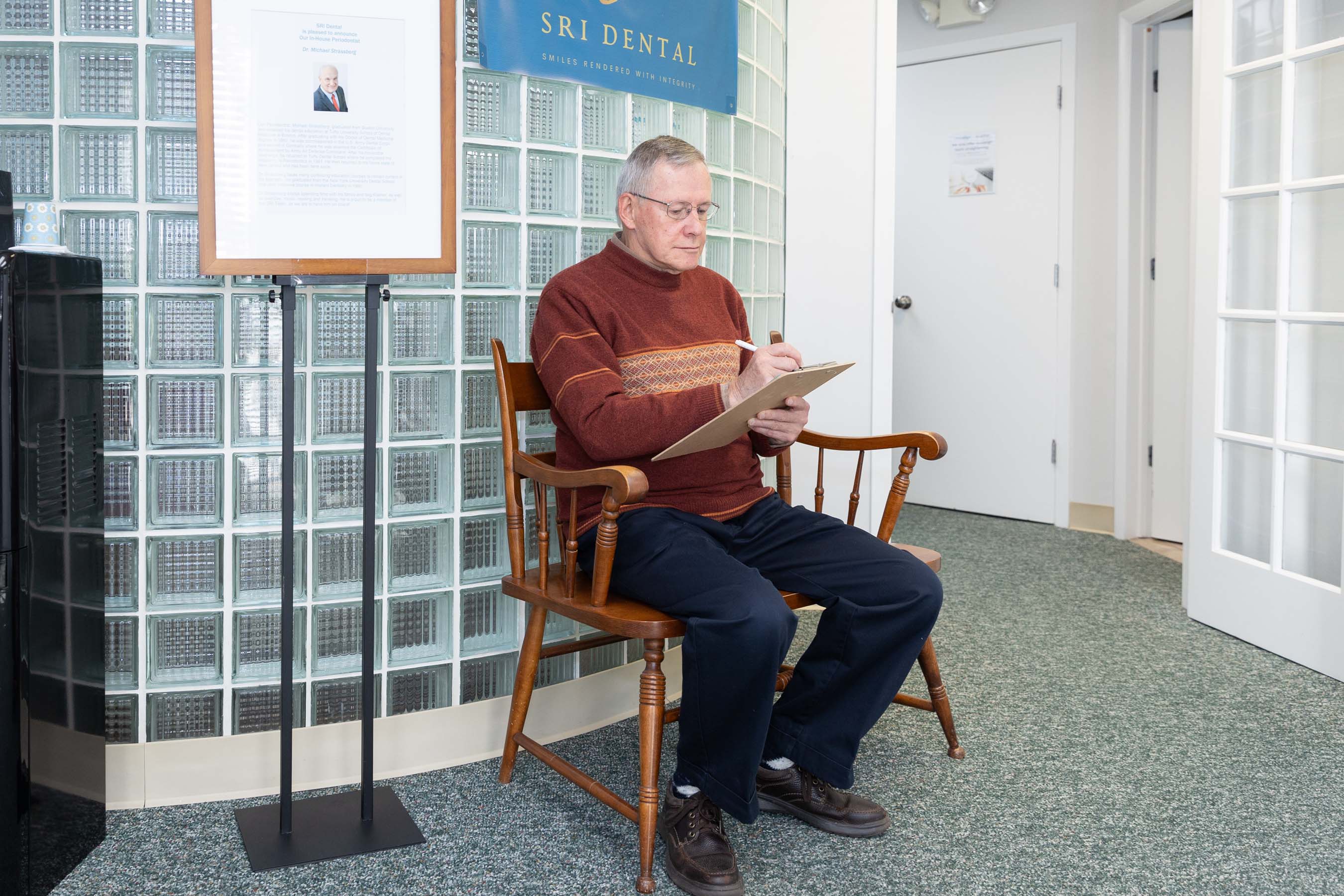 Older man in glasses sitting on wooden bench in dental office filling out a clipboard form.