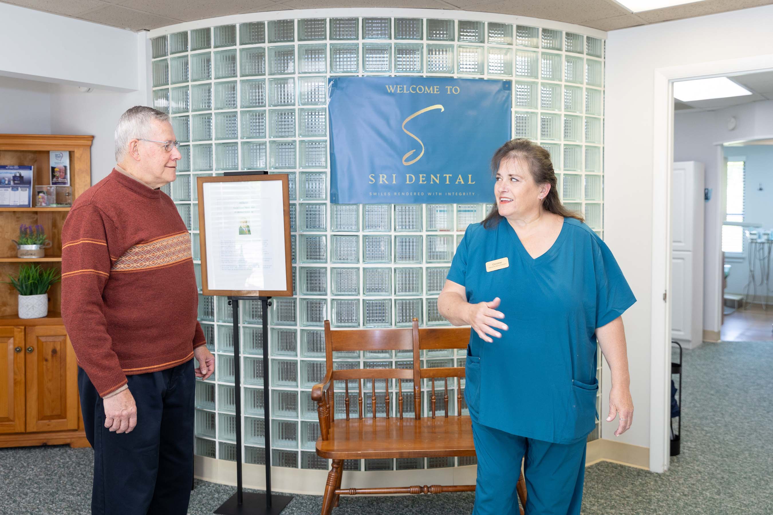 Woman in teal scrubs talking to an elderly man in a dental office waiting area.
